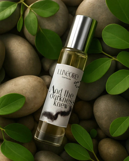 Small bottle of perfume labeled 'And the darkness knew' by Linaore on pebbles with green leaves.